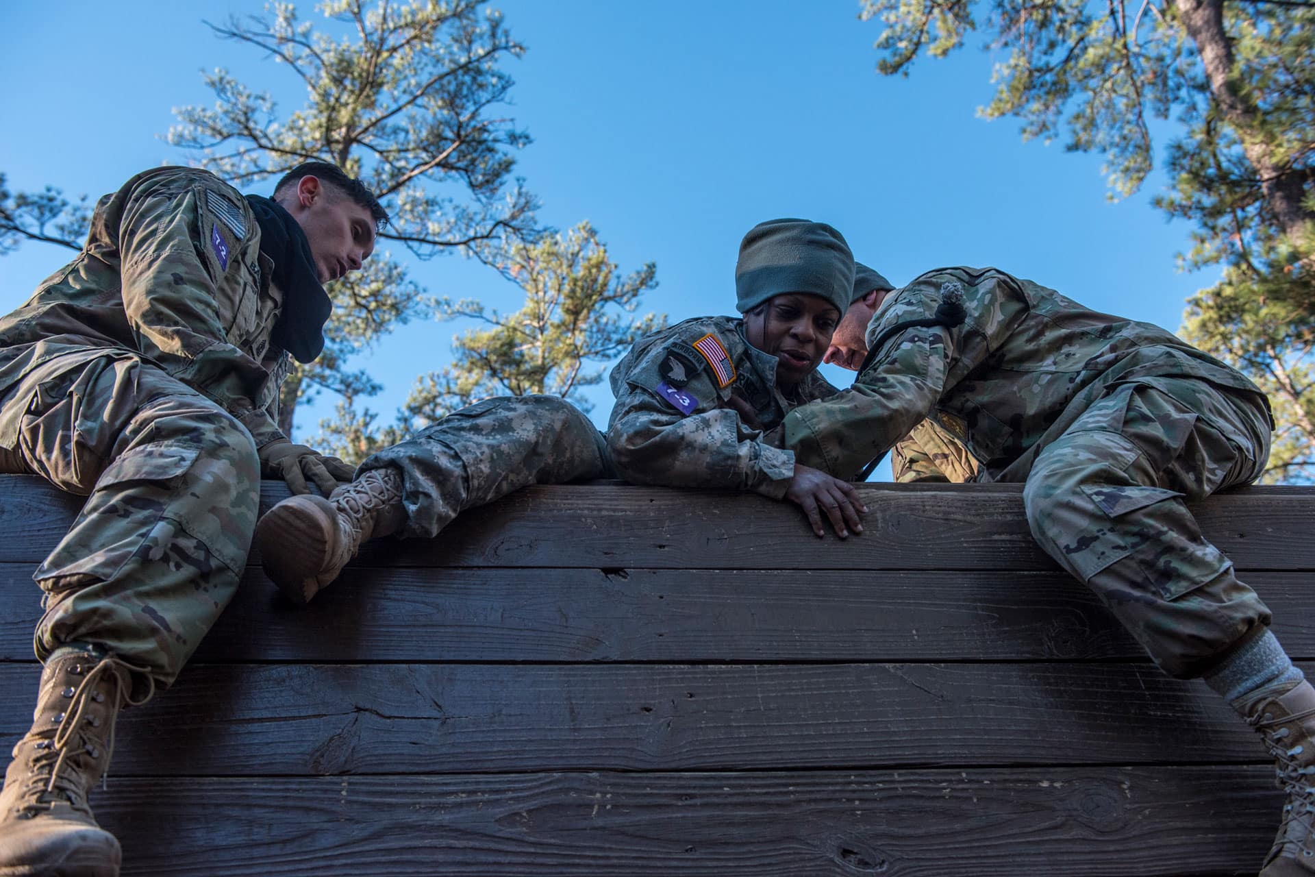 two soldiers help a fellow soldier over an obstacle wall